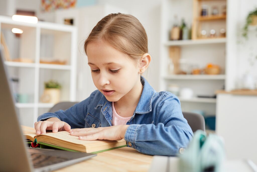 Cute Girl Reading Book to Online Teacher Warm-toned portrait of cute little girl reading text to laptop camera while studying online at home in cozy interior , copy space